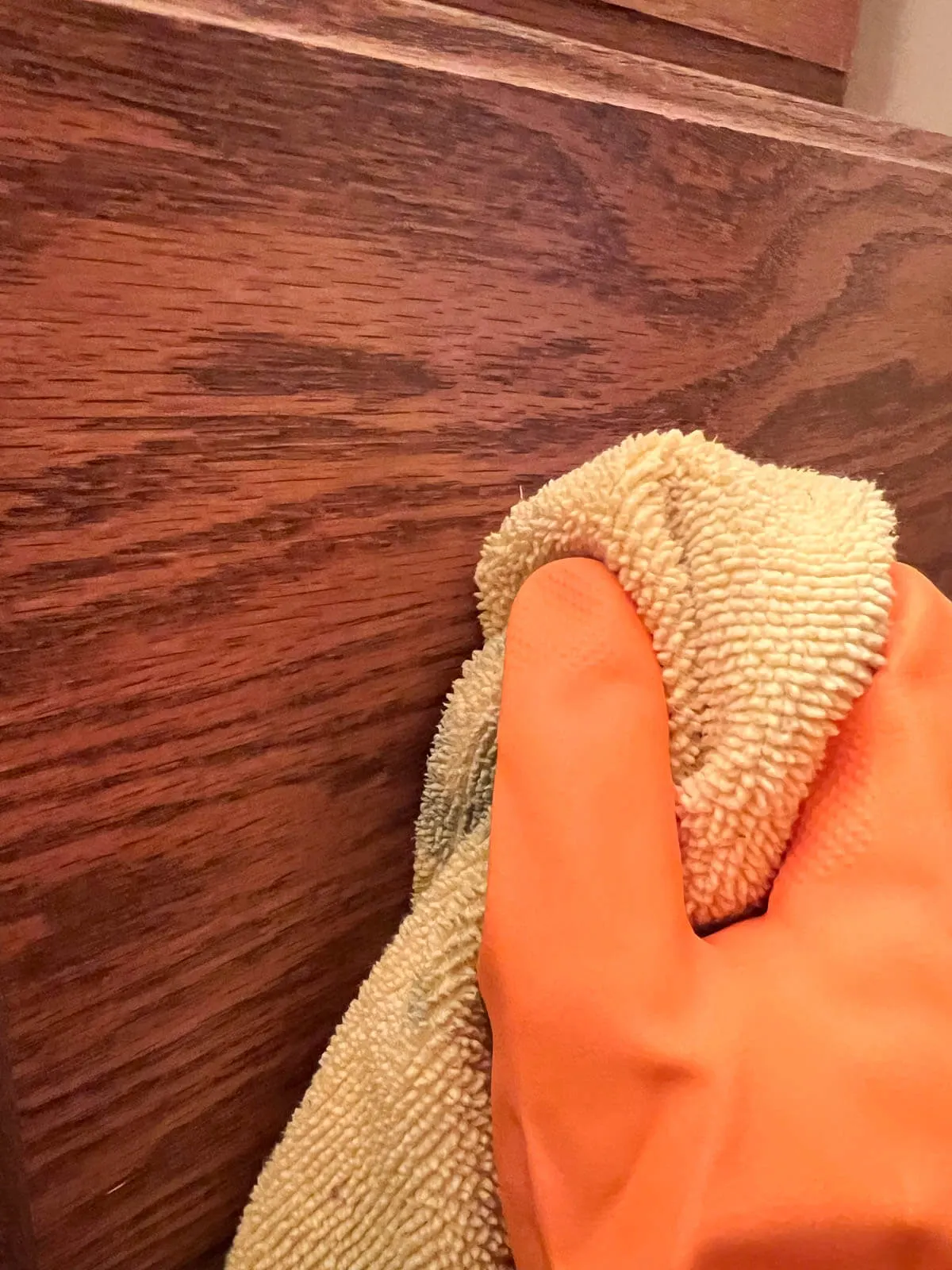 hand in orange rubber glove wiping oak cabinet with yellow rag.